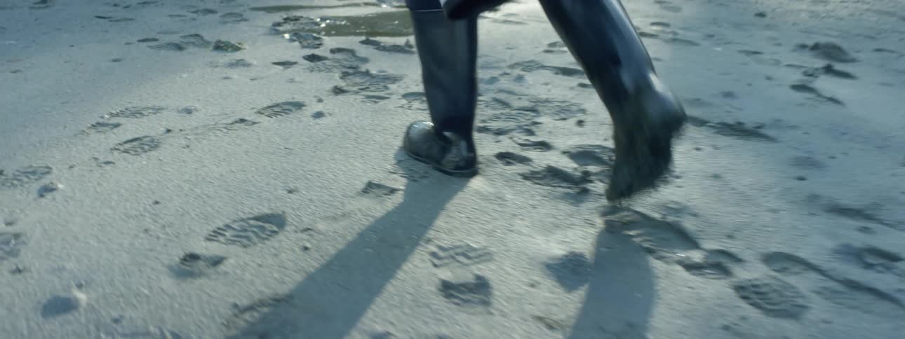 A photographer wearing boots walks on wet sand carrying a large zoom lens