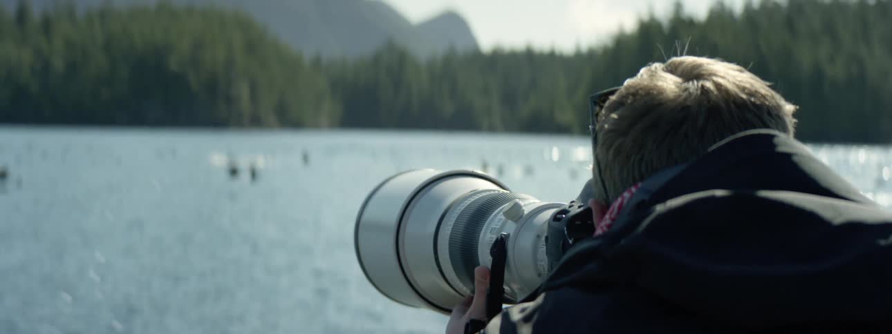 A man with a camera taking pictures of birds on the lake with trees the sky and clouds in the background