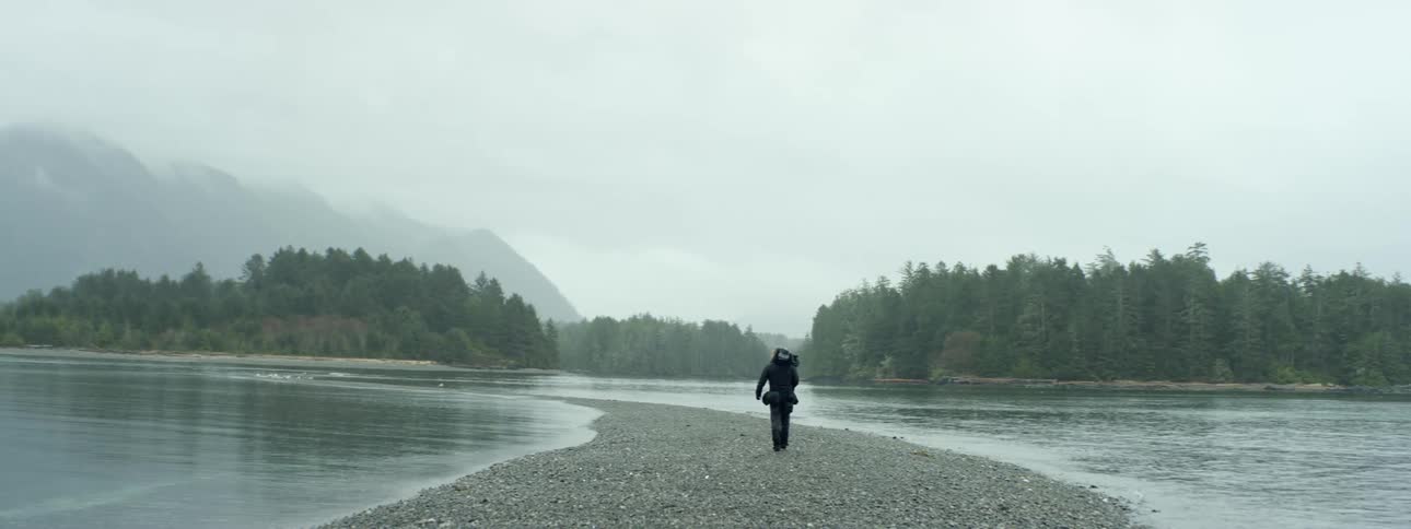 Cameraman carrying camera over his shoulder walks through dirt road between two lakes