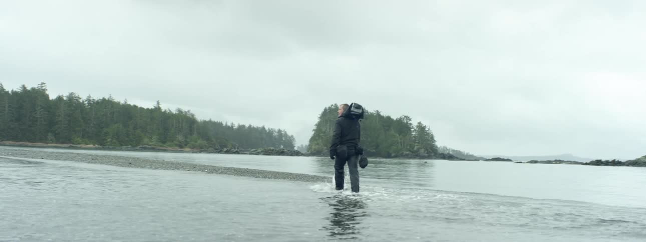 Man with black bags attached to his belt walks through water on to a strip of land with a forest of trees in front of him on a cloudy day