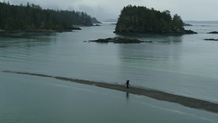 Man hikes along narrow piece of ground surrounded by water near forest covered island and shore on foggy day