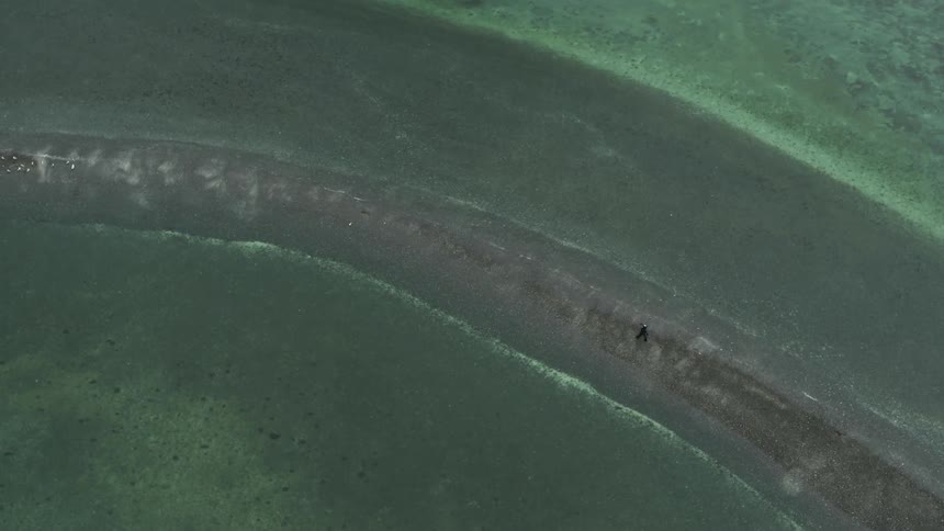 Aerial view of a man walking on a narrow sand bank through the green waters of a lake