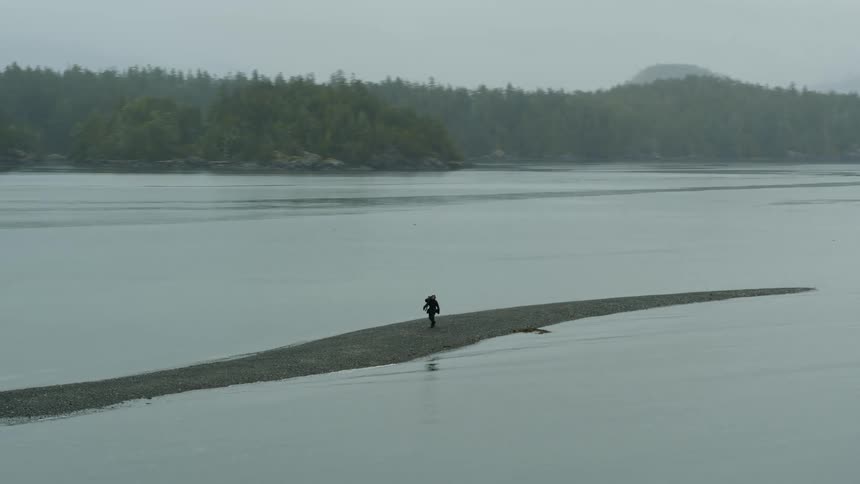 Photographer carries equipment over a sand bank in a lake on the forest near the mountains