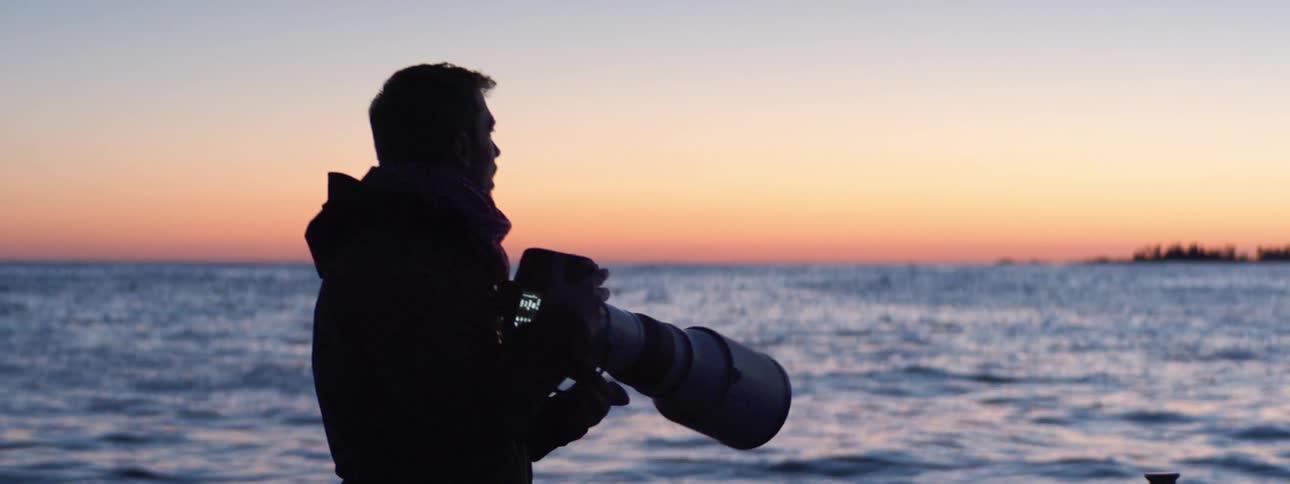 A man on a boat with a jacket and a camera with a body of water the sky and clouds in the background