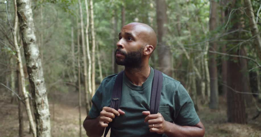 A black man gazes at his surroundings as he hikes through a forest during the day