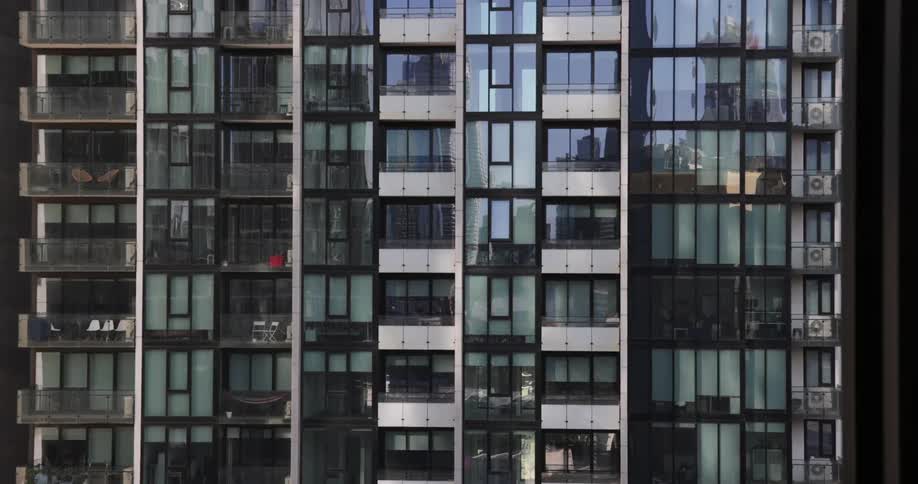 The windows of an apartment building with balconies reflect the sky and city