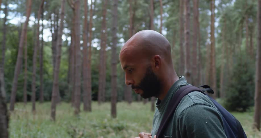 A man holds onto his backpack and looks up at the tall trees as he hikes through a forest