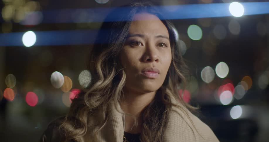 A woman stands on a city sidewalk with lights shimmering behind her at night