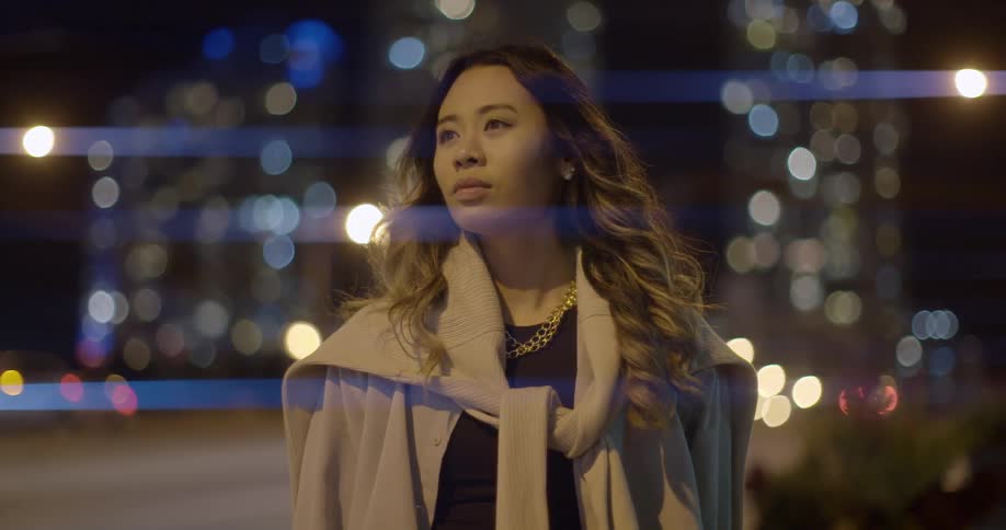 A well dressed woman stands outside in a city environment next to a road at nighttime