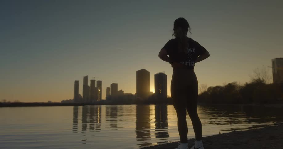 A silhouetted woman standing next to a lake with the sun setting behind skyscrapers in the distance