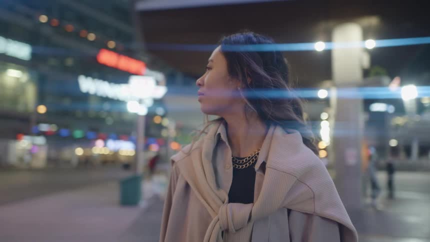 A woman stands on a city sidewalk looking around at the buildings and lights at night