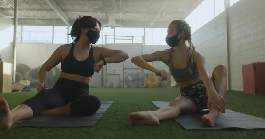 Two women sitting on yoga mats bump elbows while wearing masks