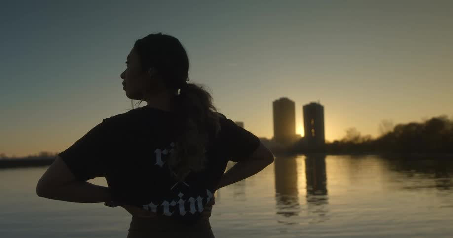 Silhouette of a young woman standing in front of a lake near a city at sunset
