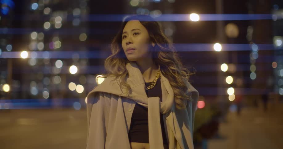 A woman stands on a downtown sidewalk and night and looks into the distance