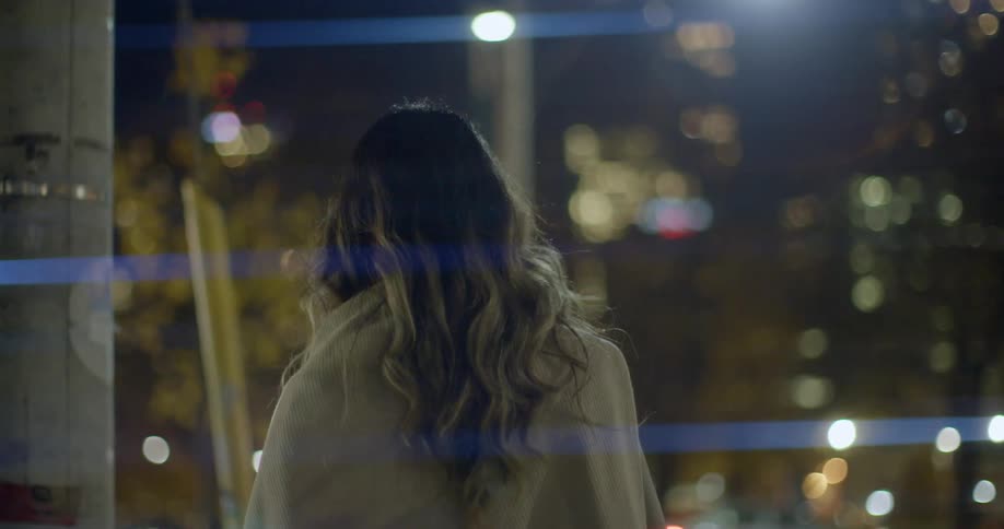A woman with long curly hair walks down a sidewalk in a busy downtown city at night