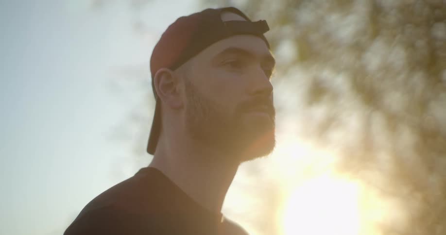 A man with a beard and wearing a baseball hat and t shirt stands outdoors at dusk
