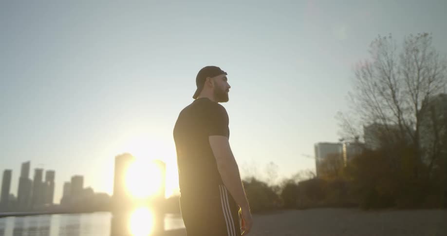 Young bearded man standing on the shore of a lake near the city skyline at sunset