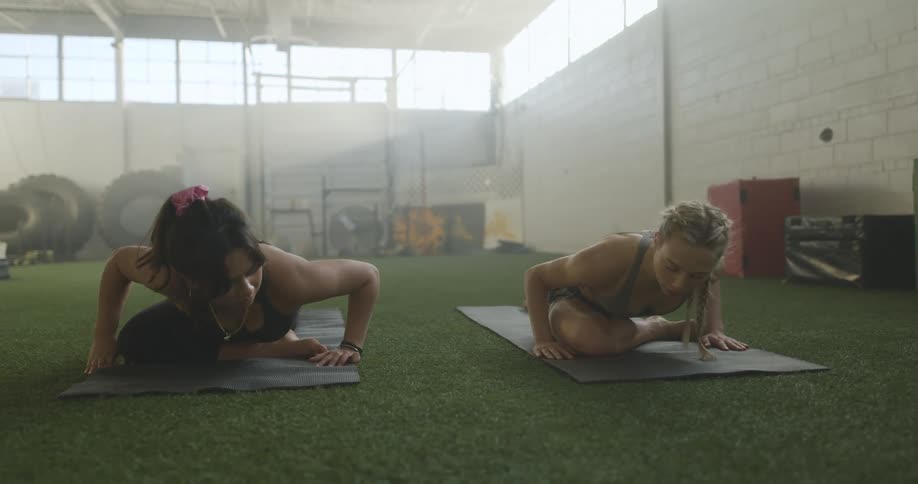 Two women are doing yoga on mats on a floor in a gym during the day