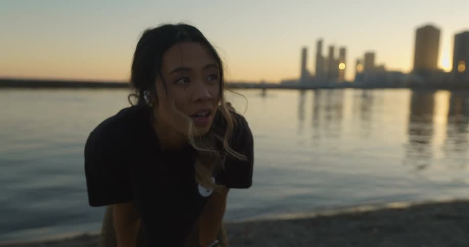 A woman with her hands on her knees catching her breath by the water looks up and laughs