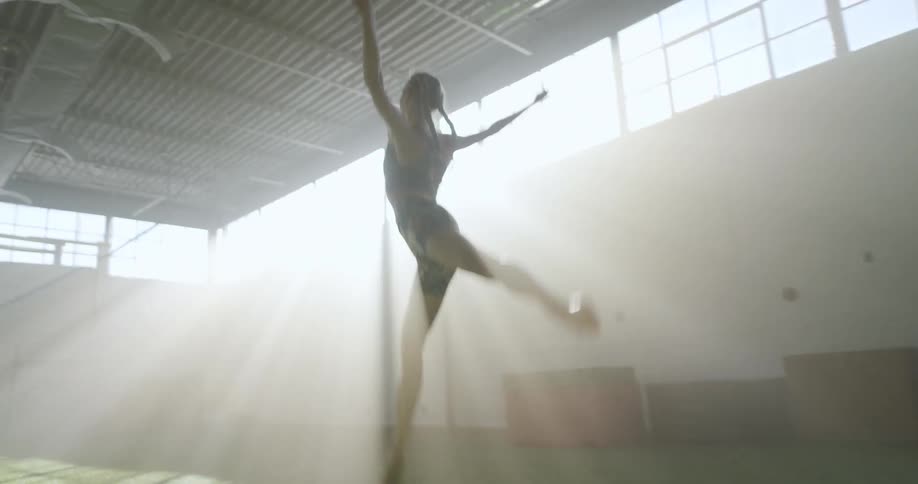 A woman gracefully runs and leaps across the turf of a gym as the sun shines through the windows