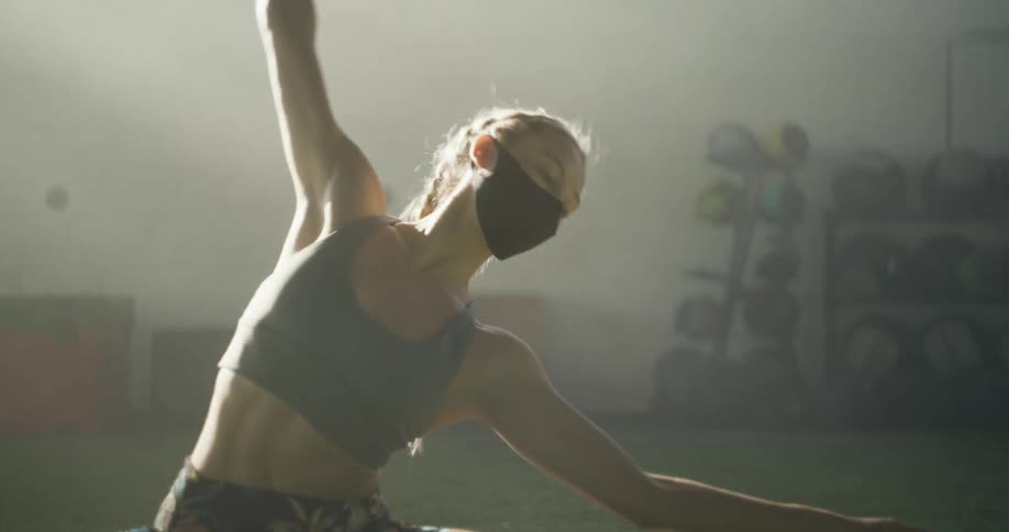 A woman in a face mask stretching in a fitness room while wearing a sports bra
