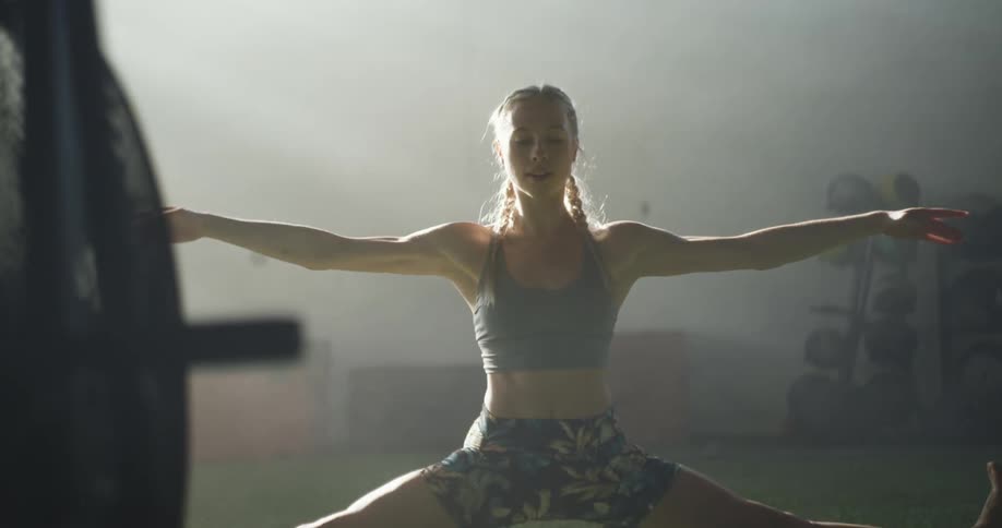 A woman in a sports bra and exercise shorts stretching and doing splits in a sunny fitness room