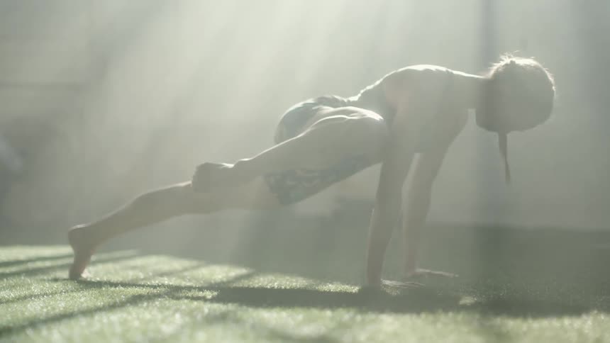 Silhouette of a young fit woman in sports clothes stretching legs over the rug of a dimly lit gym