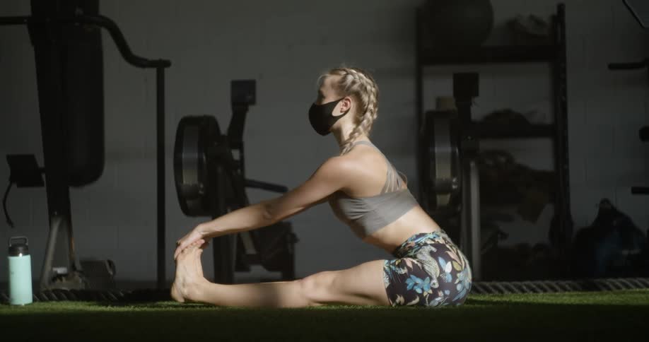 Young fit woman wearing a face mask and sports clothing stretching on the floor of a gym in the morning