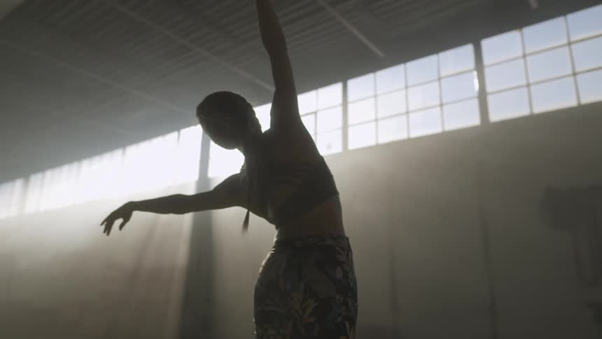 A woman takes a graceful dance pose in large dimly lit room as sunshine shines in through the windows