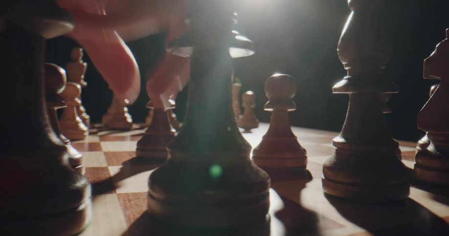 A hand moves the pawn on a wooden chessboard with a bright light shining in a dark room