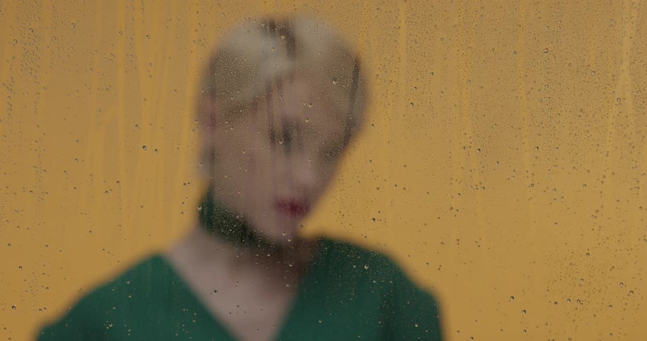 A woman with short blonde hair poses behind a pane of glass covered in water droplets