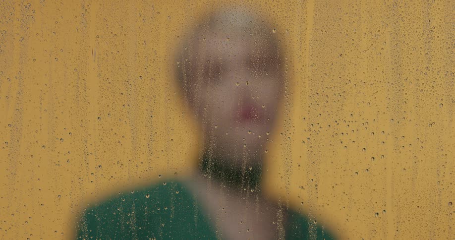 A blonde woman in a green top stands in front of a yellow wall and behind a piece of glass with water rivulets