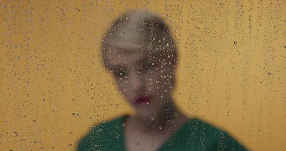 A woman in a green top walks to stand behind a glass with water droplets