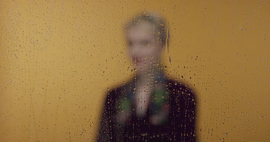 A person stands in front of a yellow wall and behind a glass pane with water droplets