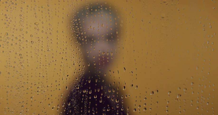 A woman standing behind rain covered glass slowly turns around to face forward