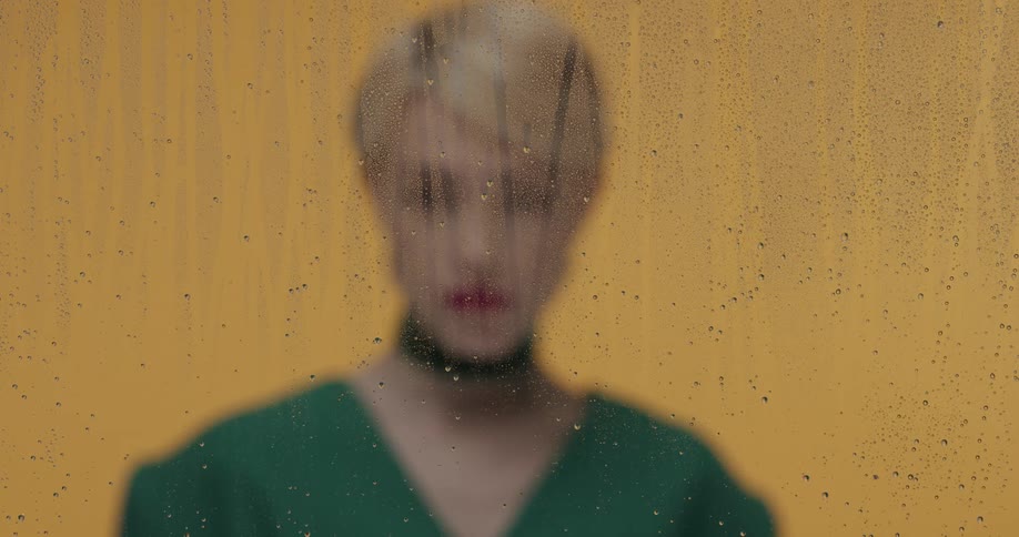 A woman with a blonde pixie cut poses behind glass that is obscured by dripping water