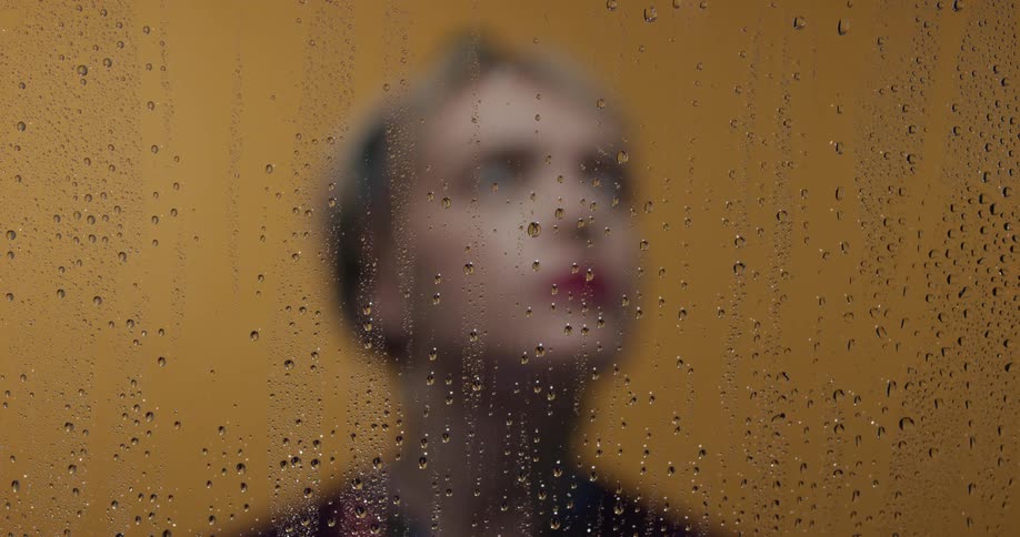 A blonde woman with red lipsticks stands behind a piece of glass with water rivulets and in front of a yellow wall