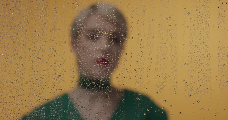 A blonde woman stands in front of a yellow wall and behind a piece of glass with water droplets