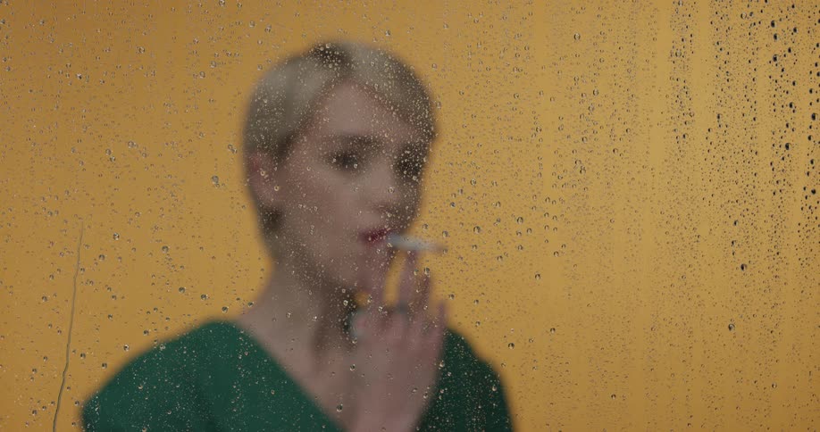 A blonde woman with short hair standing behind a wet pane of glass lights and smokes a cigarette