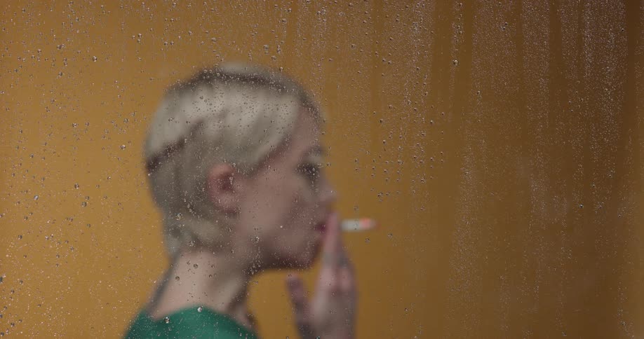 A woman is smoking by a window when its raining outside