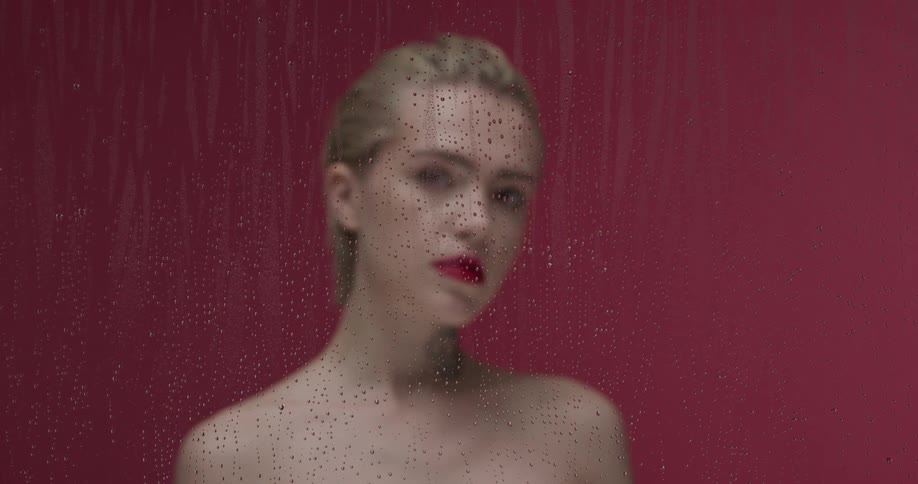 Young blonde short haired woman standing behind a wet glass pane looking at the camera