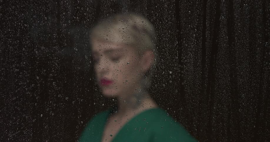 A woman in a green top is smoking a cigarette while standing behind a pane of glass with water droplets