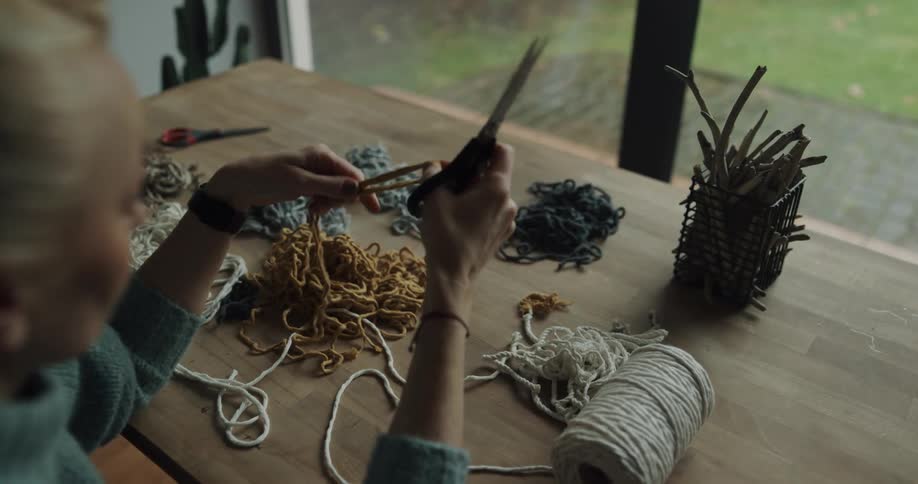 A woman is cutting yarn while sitting next to a window indoors