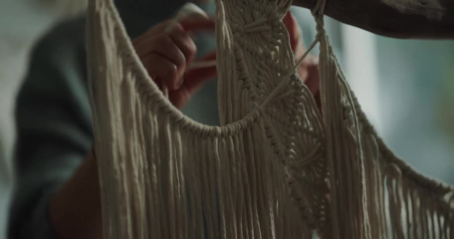 Woman knots cords on macrame project hanging from wooden branch