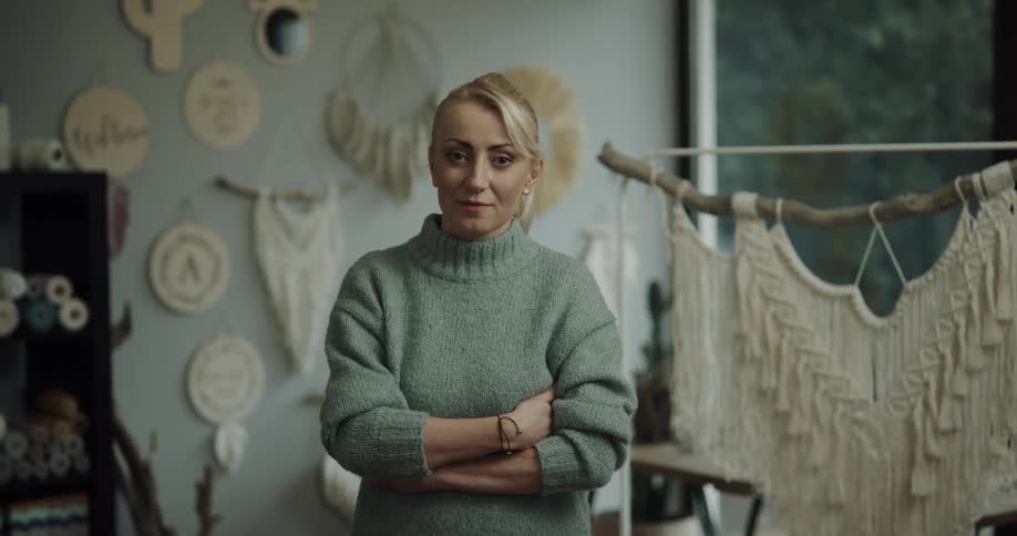 A woman stands and poses for the camera with many knitted art pieces hanging behind her