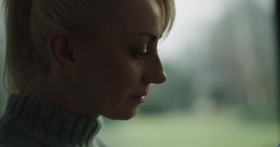 A young woman in a sweater slides her finger across the screen of her smartphone while standing near a window indoors during the day