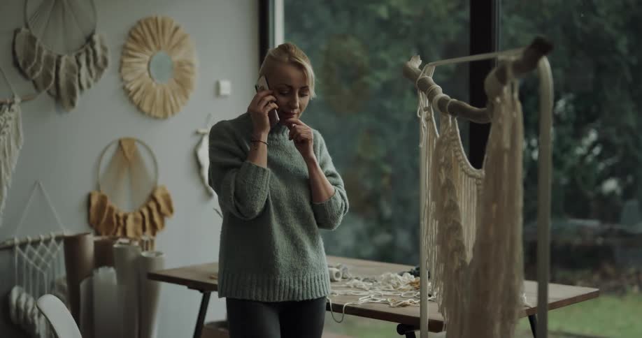 A young woman talks on a smartphone while standing in front of a table covered in yarn and string near a window indoors during the day