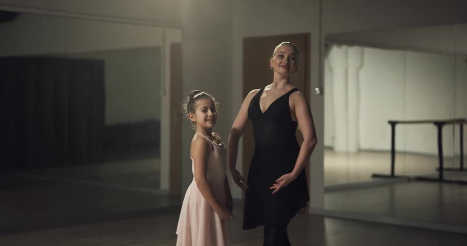 A young girl and a woman ballet dancers stand together smiling in a studio with mirrored walls