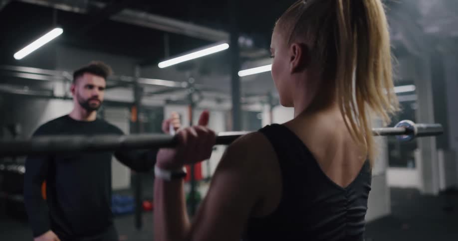 A woman lifting weights in an indoor gym while a man coaches her