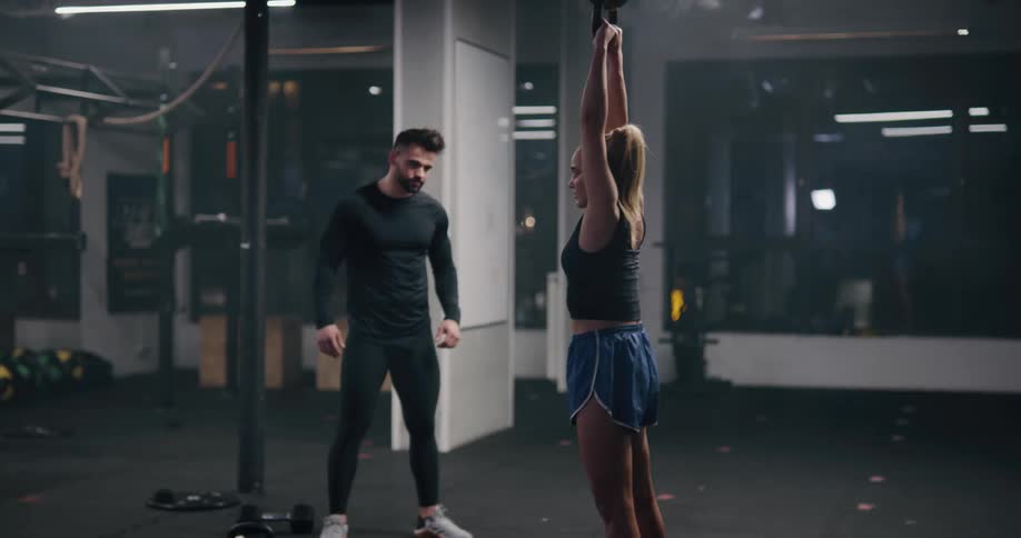 A trainer watches as a young woman swings a kettle bell weight above her head repeatedly